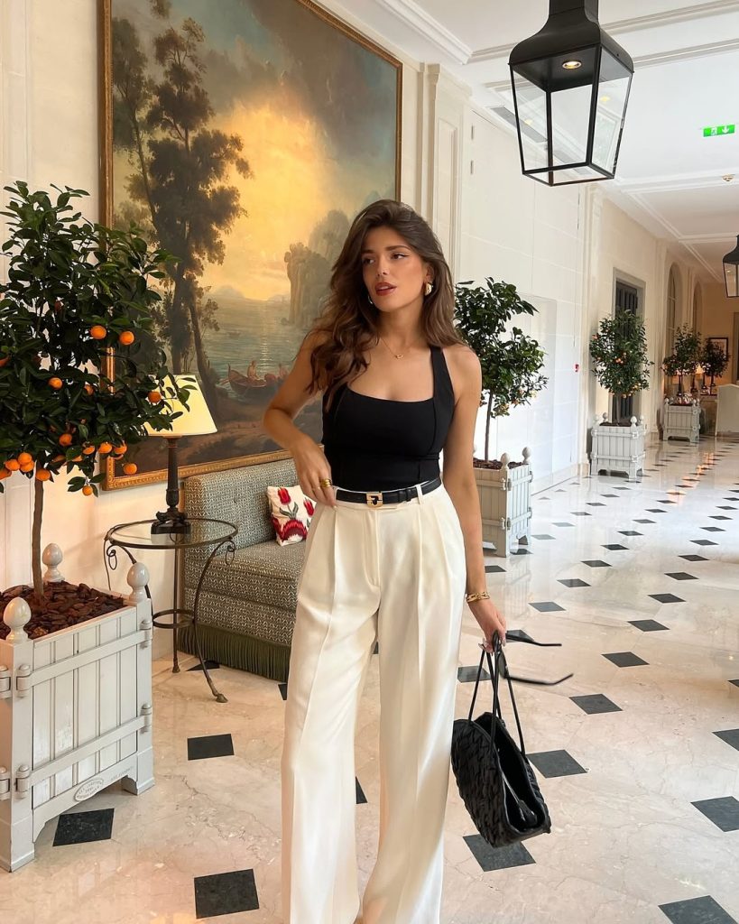 Old money summer outfit with fitted black tank top, cream wide-leg trousers, designer belt with gold buckle, gold jewelry and quilted black shoulder bag
