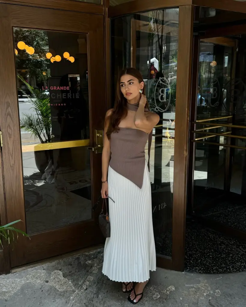 classy summer date night outfit with brown asymmetric bandeau top, matching neck scarf, white pleated maxi skirt, minimal black sandals and subtle brown handbag 