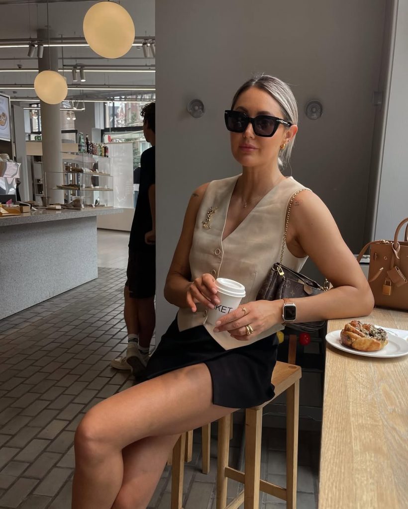 Elegant old money summer outfit with beige sleeveless button-front vest, black mini skirt, designer monogram crossbody bag, black oversized sunglasses and gold jewelry