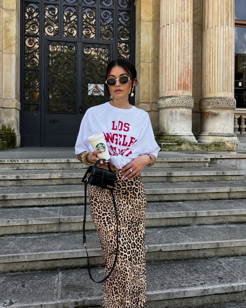 Casual summer outfit with white graphic sweatshirt, leopard print maxi skirt, round sunglasses, gold statement jewelry and small black crossbody bag