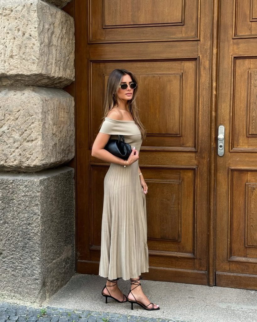 Old money summer outfit with beige off-shoulder pleated maxi dress, strappy black heeled sandals, black leather clutch, slim gold bracelets and round sunglasses
