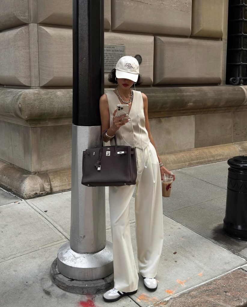 Old money summer outfit with cream sleeveless button-front vest, matching cream silk trousers, white baseball cap, chunky silver necklace, black structured designer bag and white sneakers

