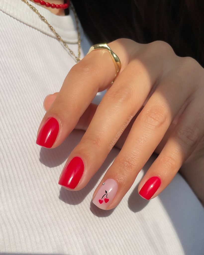 Short square nails with glossy raspberry-red polish and one accent nail with red cherry design on sheer pink base
