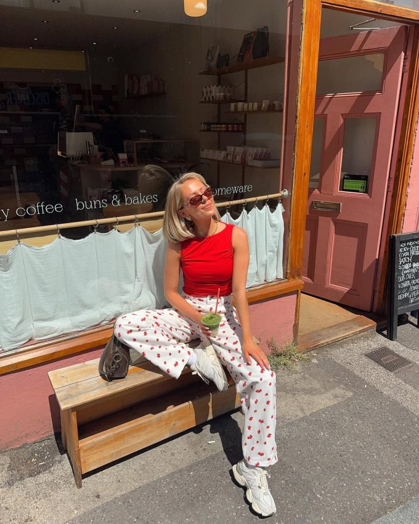 Casual summer outfit with bright red sleeveless top, white jeans with cherry print pattern, tinted sunglasses, gray handbag and white chunky sneakers
