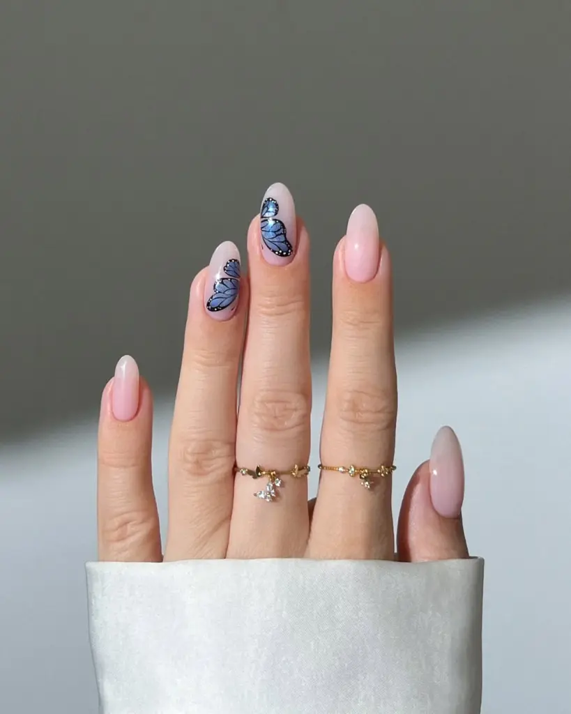 Classy June nails with almond shape featuring pale pink base with blue butterfly nail art accents