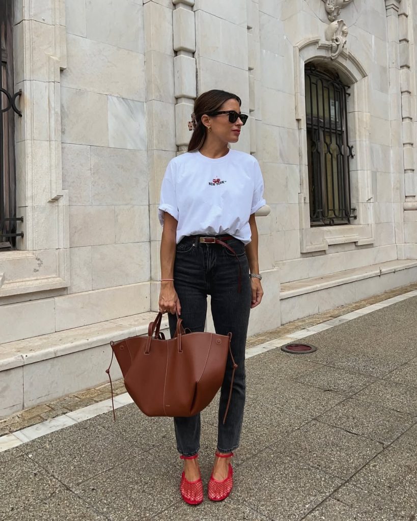 Casual summer outfit with white graphic t-shirt, dark gray straight-leg jeans, red skinny belt, oversized brown leather tote bag and bright red woven flats
