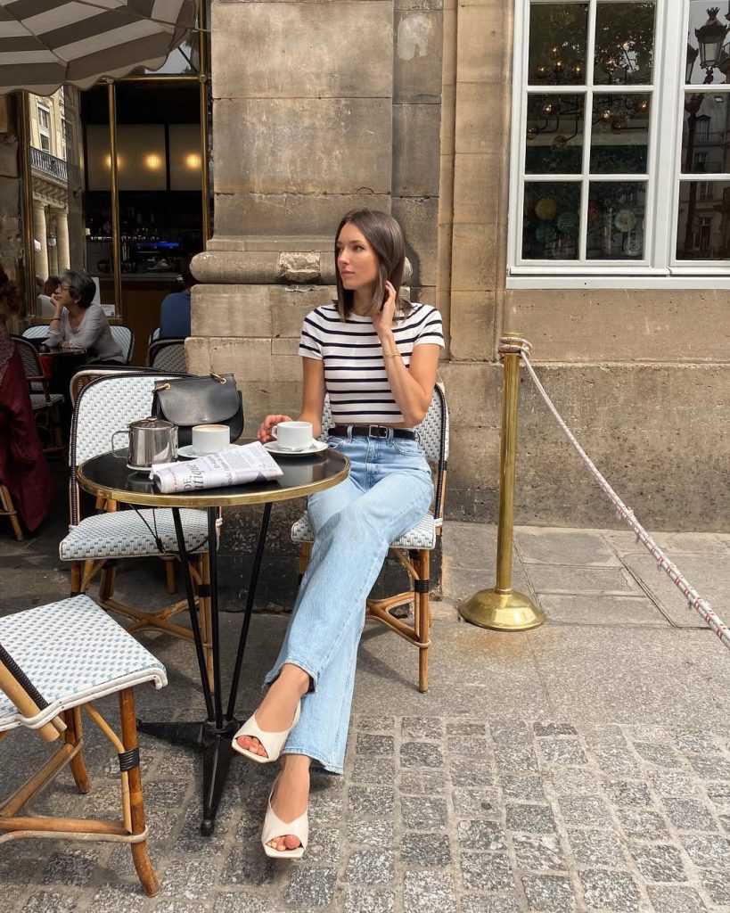Parisian summer outfit with white and navy striped t-shirt, light wash straight leg jeans, brown leather belt, white sandals and black structured handbag