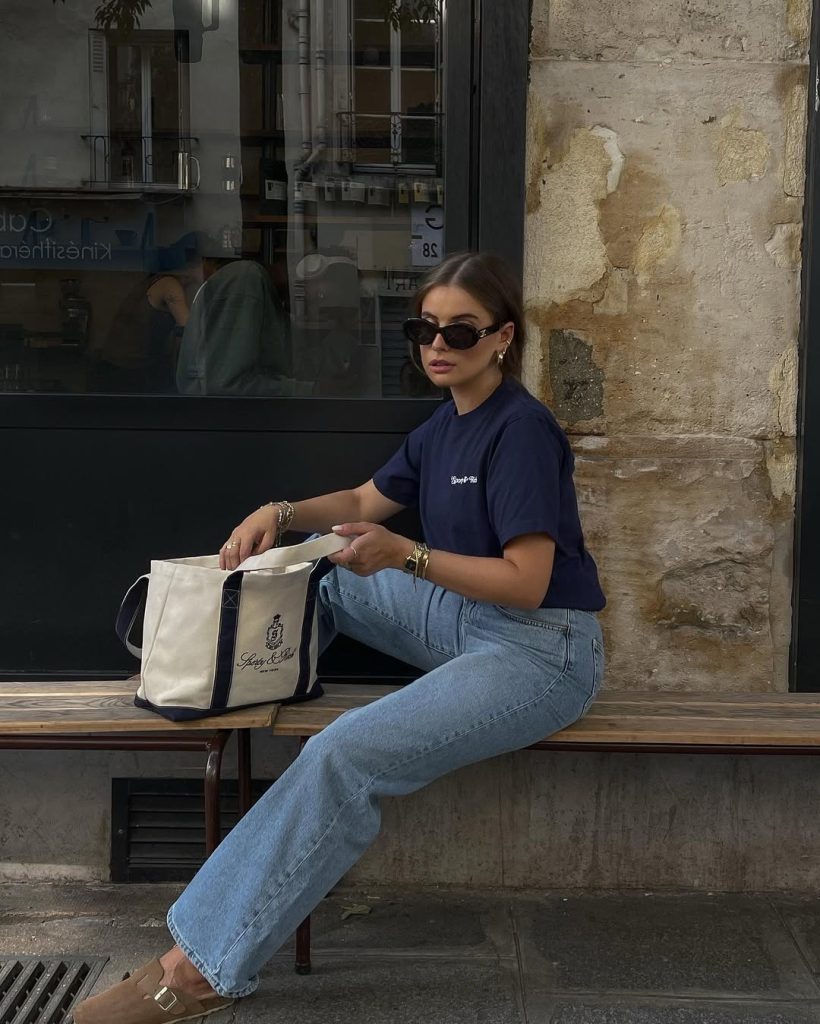 Casual summer outfit with dark blue graphic t-shirt, light wash straight-leg jeans, black cat-eye sunglasses, gold jewelry and cream canvas tote bag
