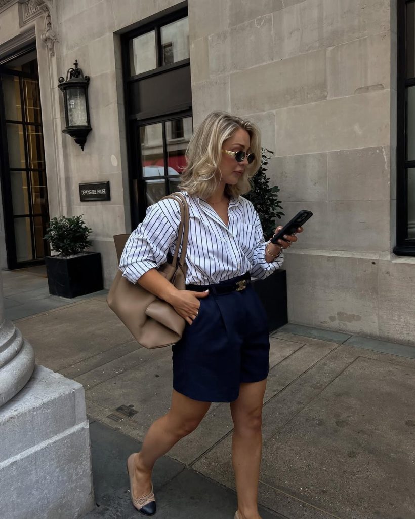 Old money summer outfit with white and navy striped button-down shirt, navy tailored shorts, designer belt, beige structured tote bag and two-tone ballet flats