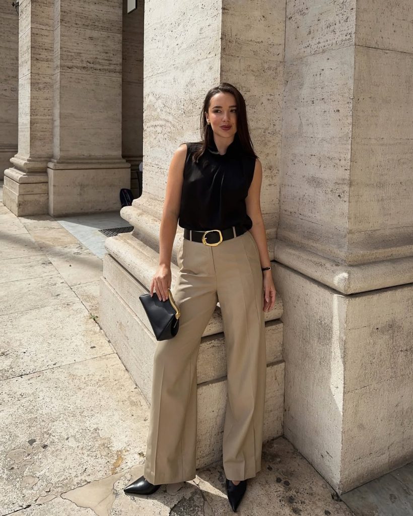 Elegant summer work outfit with sleeveless black tie-neck blouse, beige wide-leg trousers, statement gold belt and black pointed heels