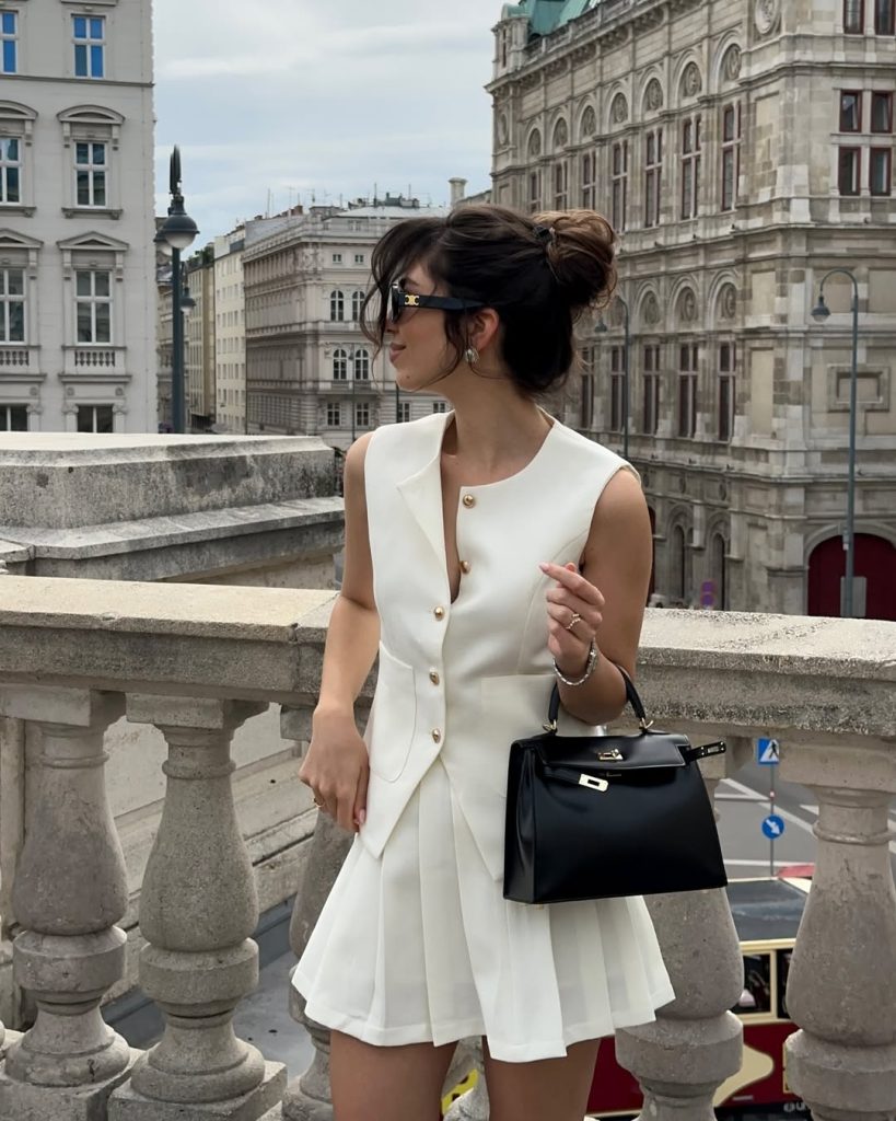 Old money summer outfit with white sleeveless button-front vest, matching white pleated mini skirt, black structured designer handbag, and gold sunglasses