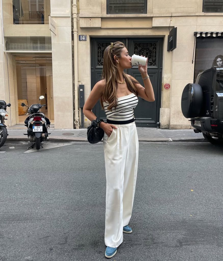 Casual summer outfit with white and black striped strapless top, cream wide-leg trousers, chunky gold necklace, black clutch bag and blue platform loafers