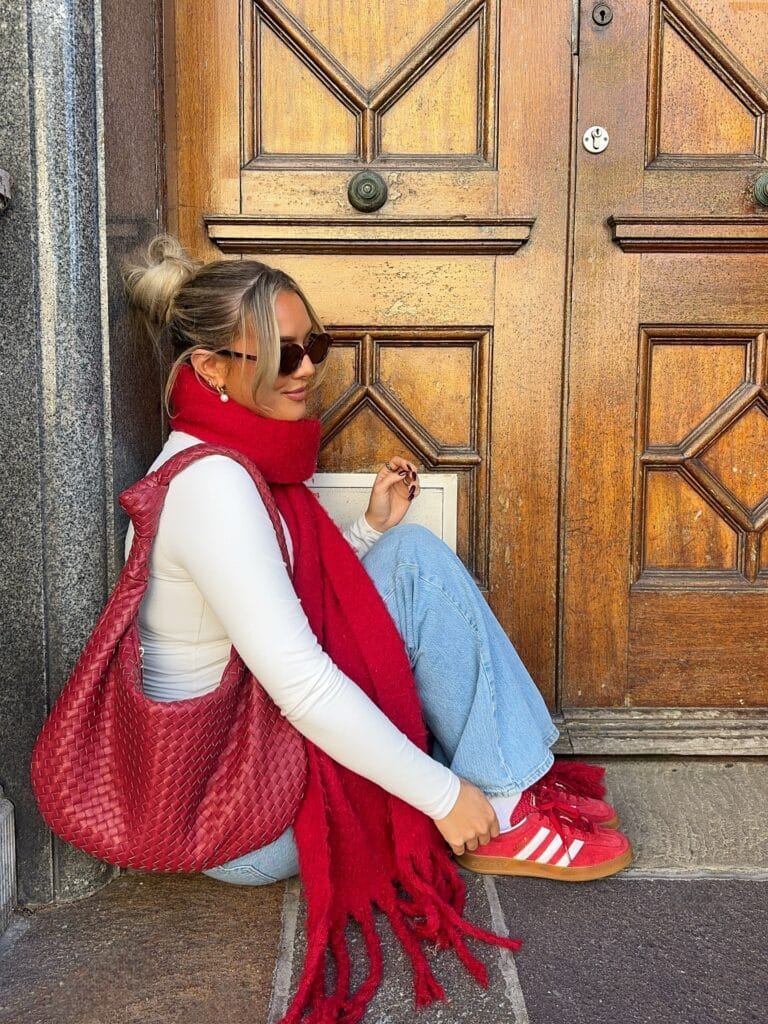 Cozy fall outfit with white top, light-wash jeans, oversized red scarf, red sneakers, and woven bag.
