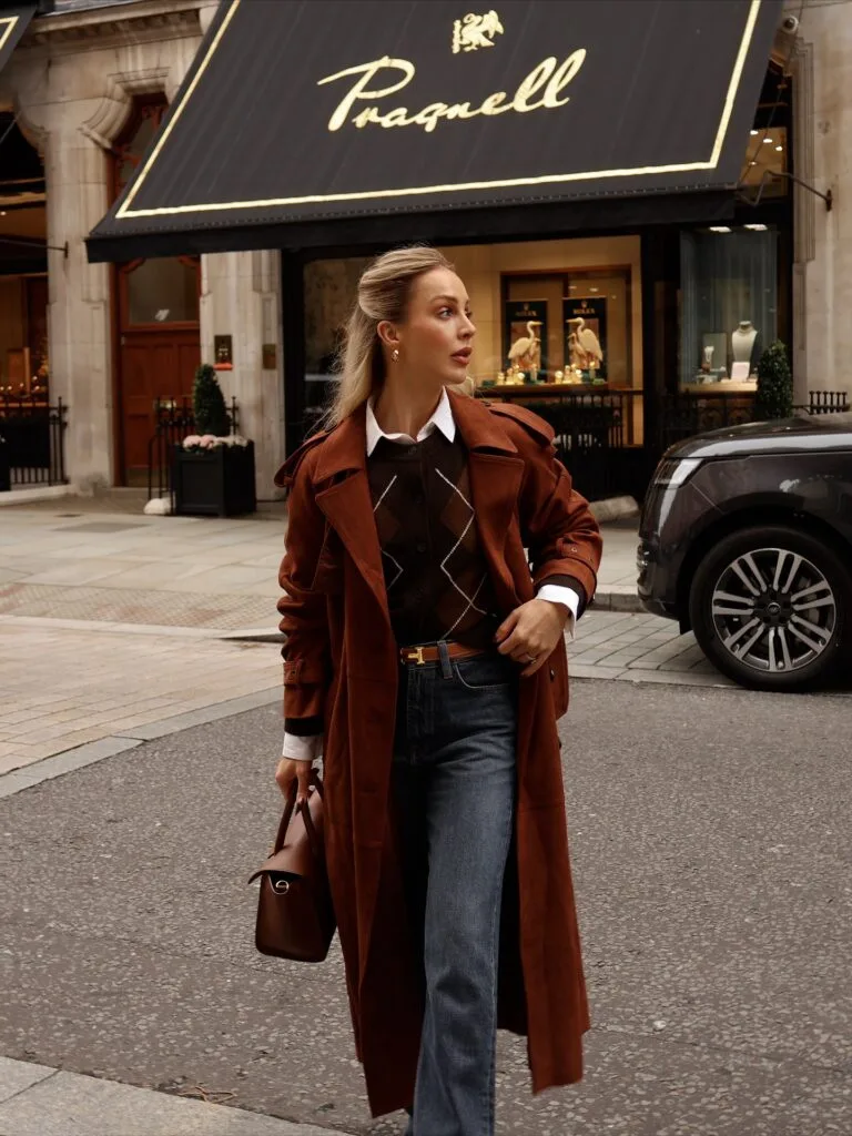 old money fall outfit with rust suede trench coat, brown argyle knit, white shirt and blue jeans