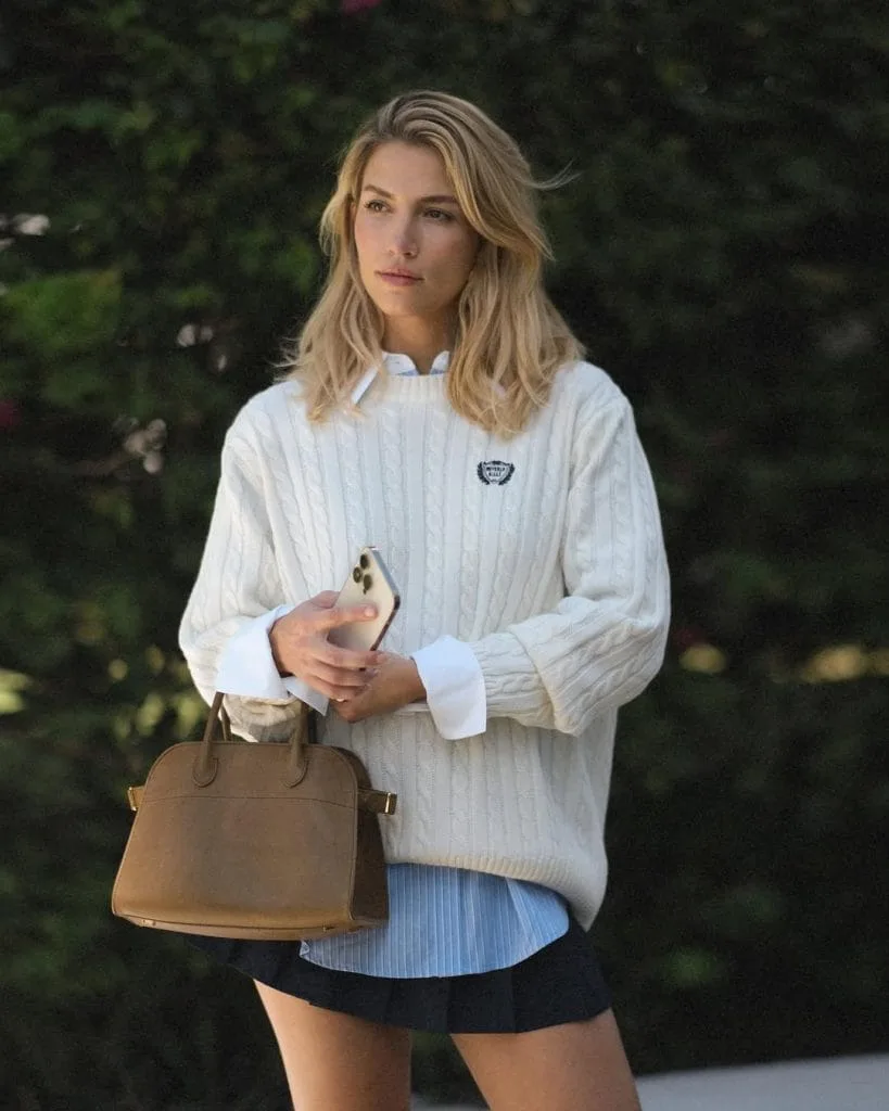 Classic old money fall outfit with cream cable knit sweater, light blue striped button-down shirt, navy mini skirt, and structured tan leather handbag
