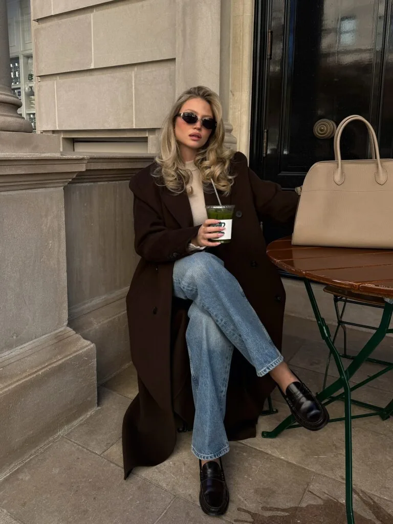 Casual autumn outfit with long dark brown coat, beige knit, light wash jeans, brown loafers, and tan tote
