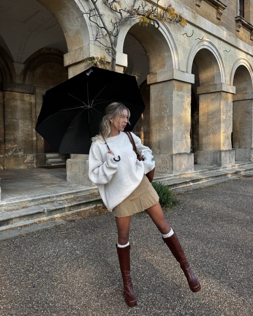 Casual fall outfit with cream oversized sweater, khaki pleated mini skirt, and brown knee-high boots