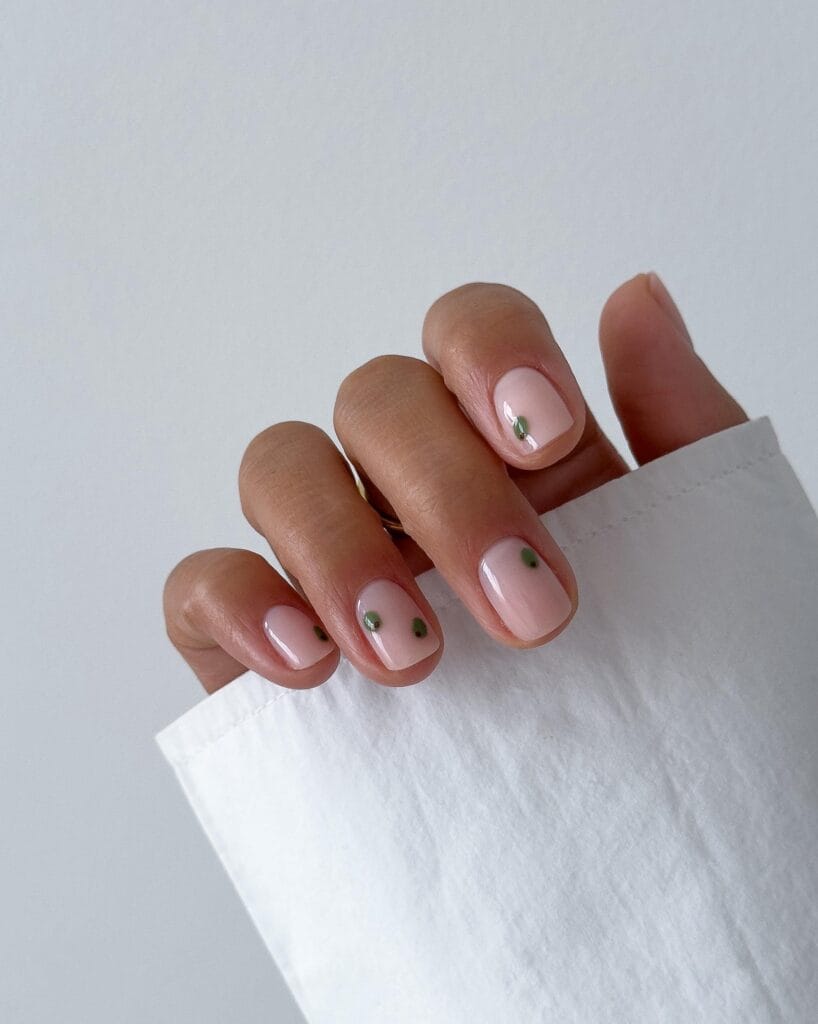 Short square pale pink nails with tiny green olive designs and reddish-brown centers.
