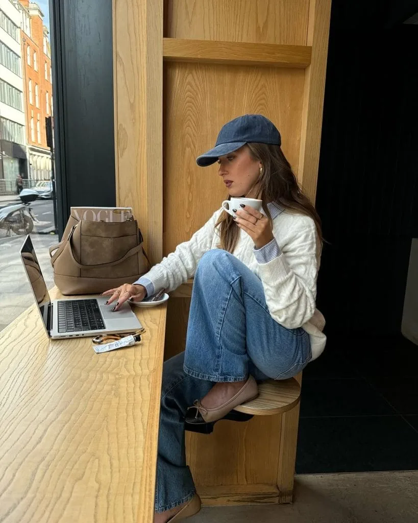 Casual fall outfit with white cable knit sweater, striped collar shirt, medium wash jeans, navy baseball cap, brown cap toe flats