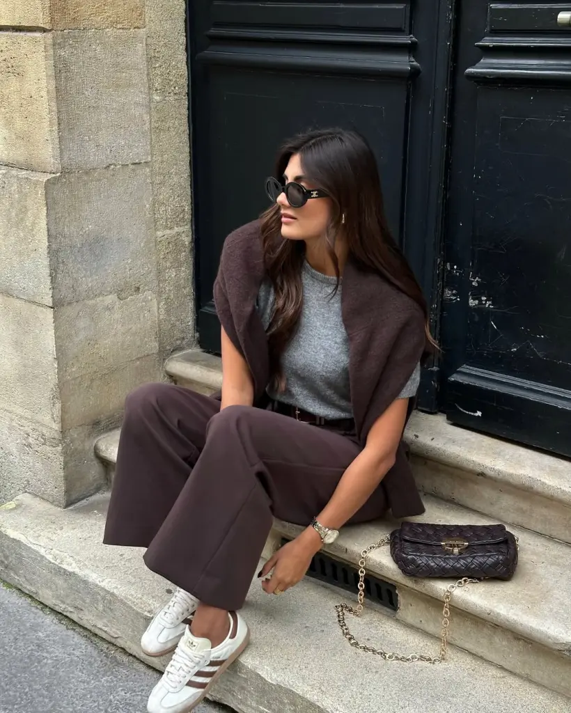 fall outfit with brown sweater worn over shoulders, grey knit sweater, brown wide-leg trousers, white sneakers and black quilted chain bag
