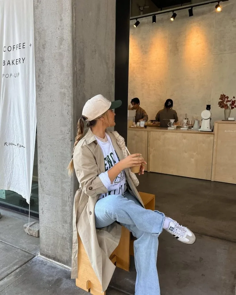 Casual fall outfit with beige trench coat, white graphic sweatshirt, light wash jeans, white baseball cap, white sneakers
