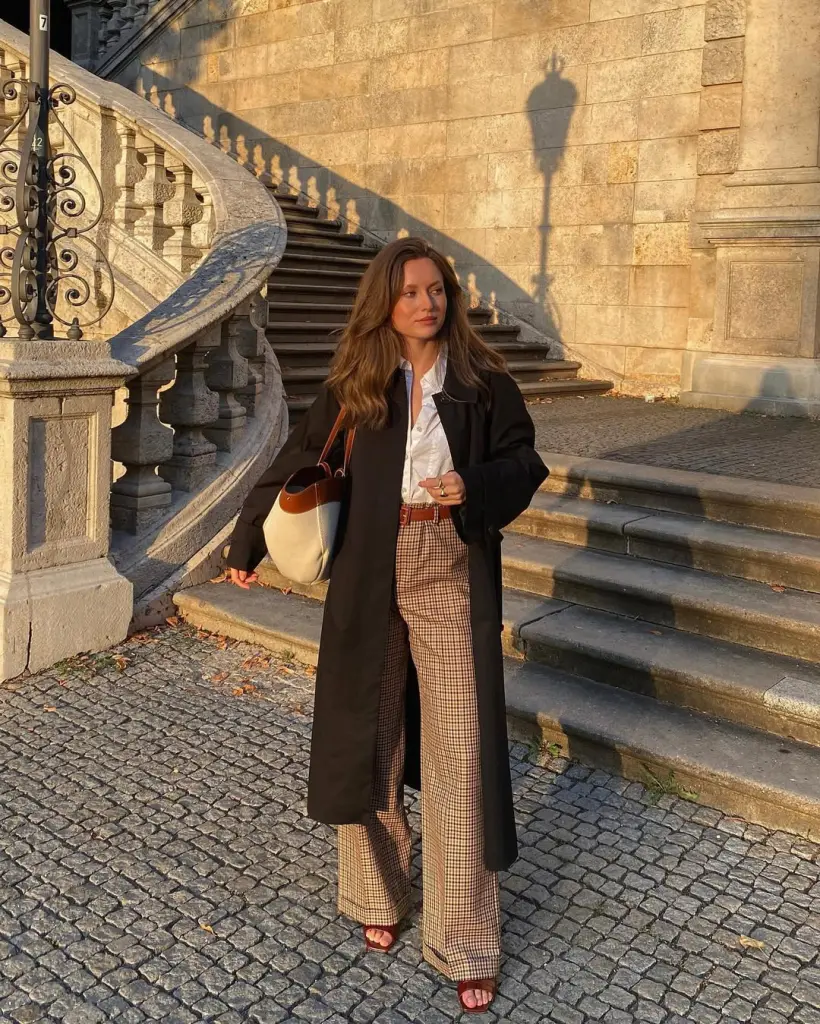 Elegant fall outfit with long black coat, white button-down shirt, brown check wide-leg trousers, brown strappy sandals and cream and brown shoulder bag
