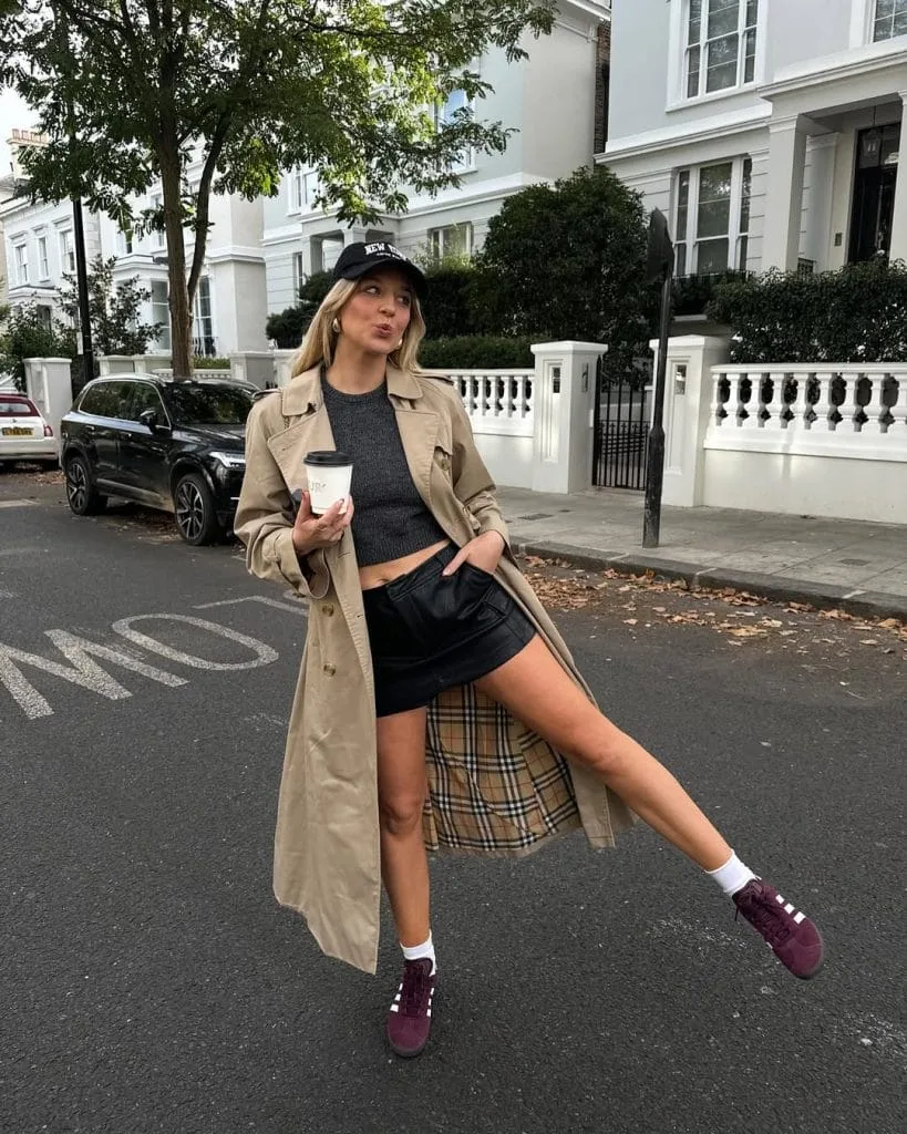 Casual fall outfit with beige trench coat, gray cropped sweater, black leather mini skirt, burgundy sneakers
