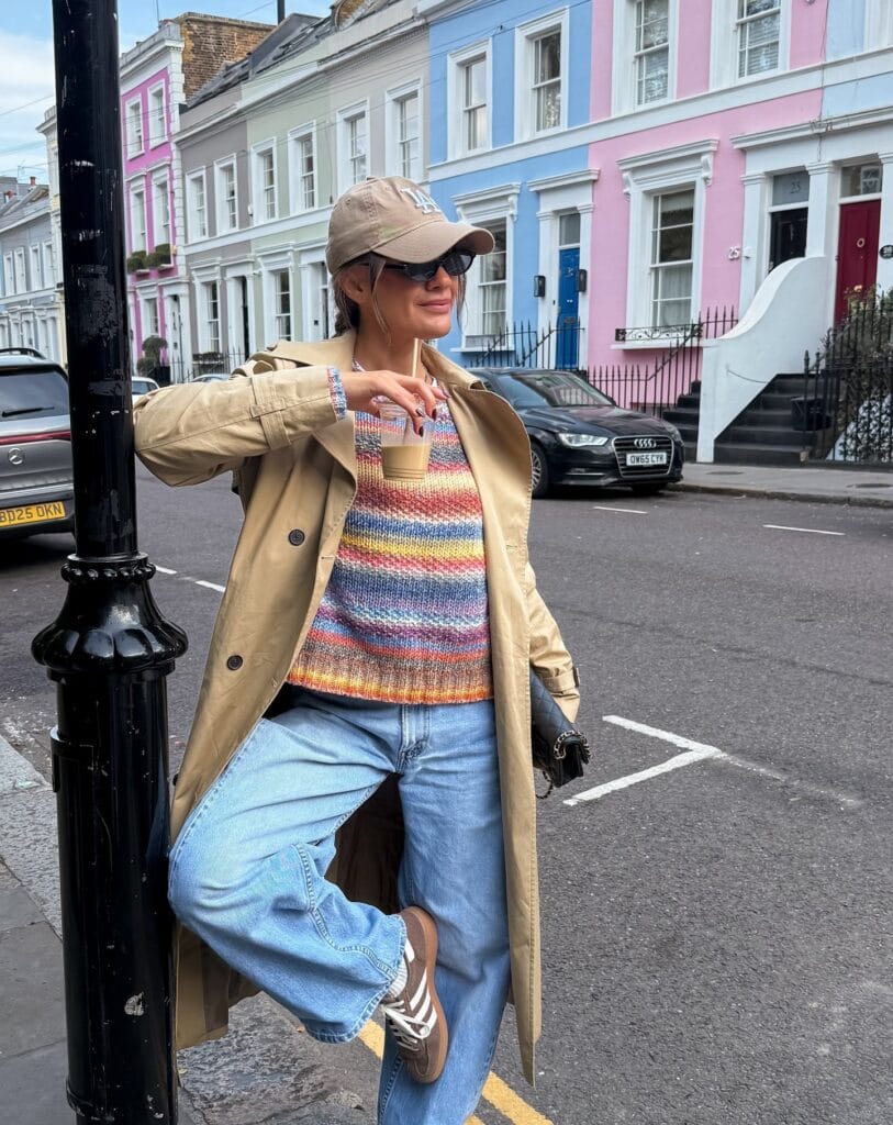 Fall outfit with beige trench coat, striped sweater, light-wash jeans, cap, and brown sneakers.
