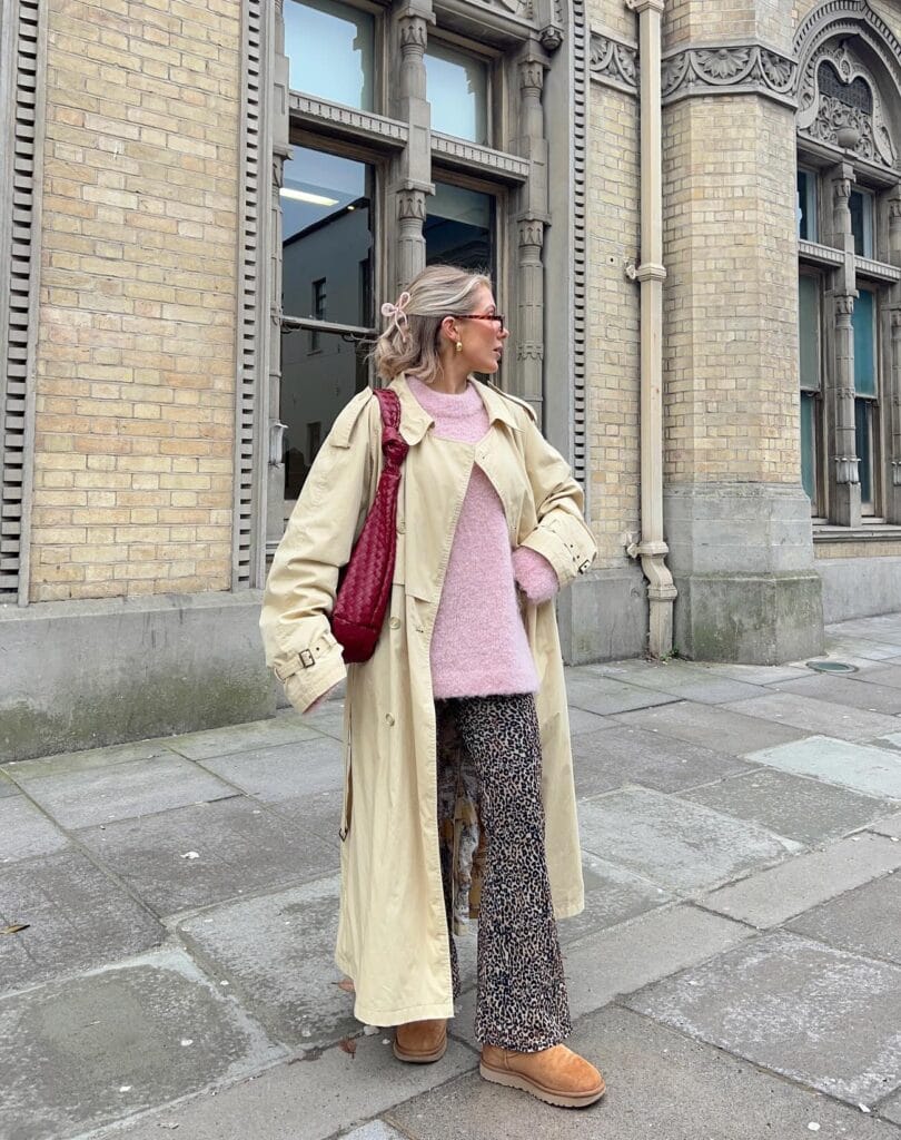 Cozy fall outfit with pink sweater, leopard-print pants, beige trench coat, tan UGGs, and burgundy bag.
