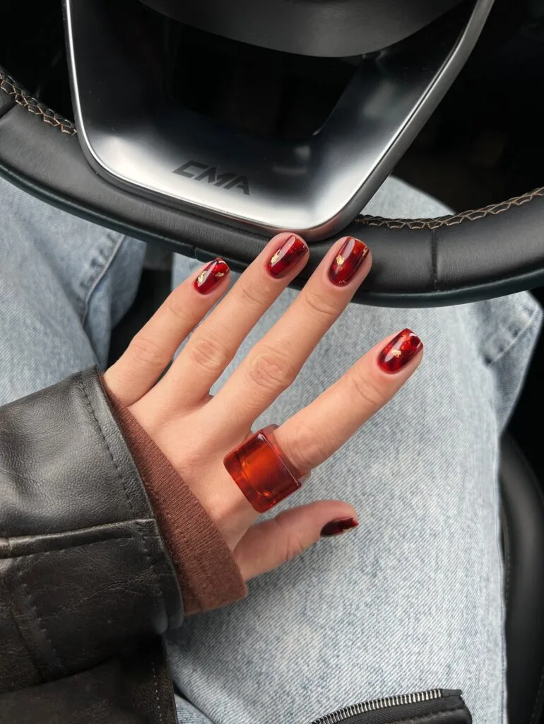 Short fall nails with translucent cherry-red tortoiseshell and small gold foil accents.
