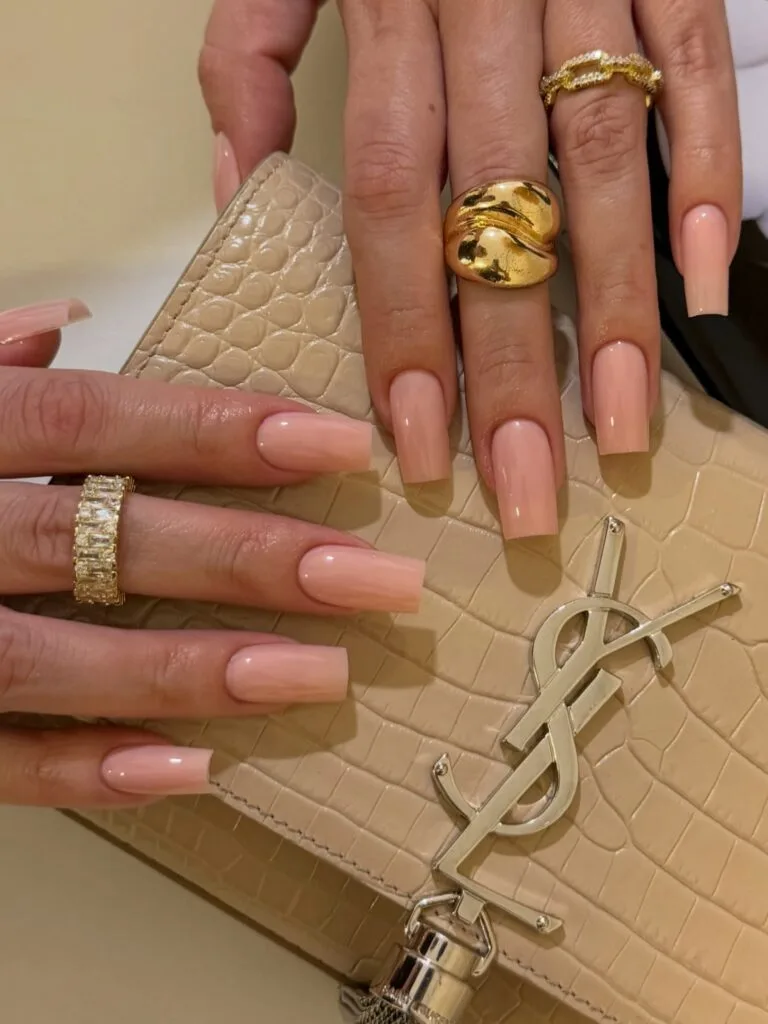 Long square nails in glossy peach nude, styled with chunky gold rings.