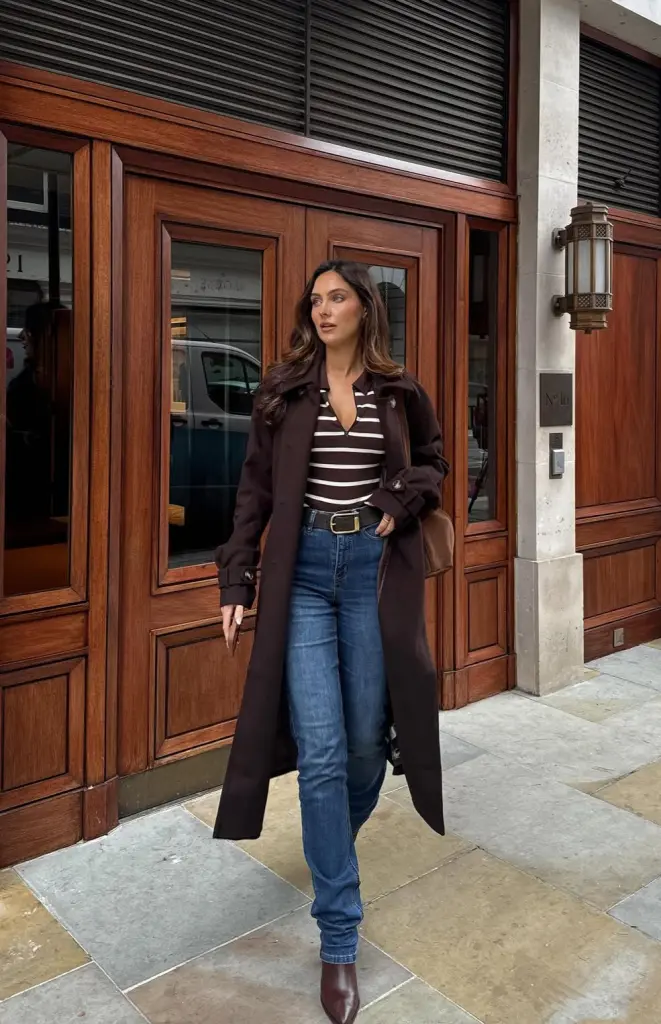 Chic fall outfit with brown long trench coat, brown and white striped top, high-waisted jeans, brown ankle boots and black belt
