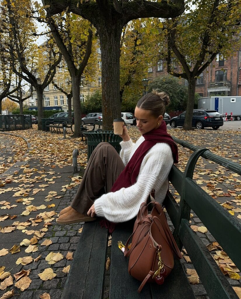 Cozy fall outfit with white knit sweater, soft brown pants, burgundy scarf, tan UGGs, and brown bag.
