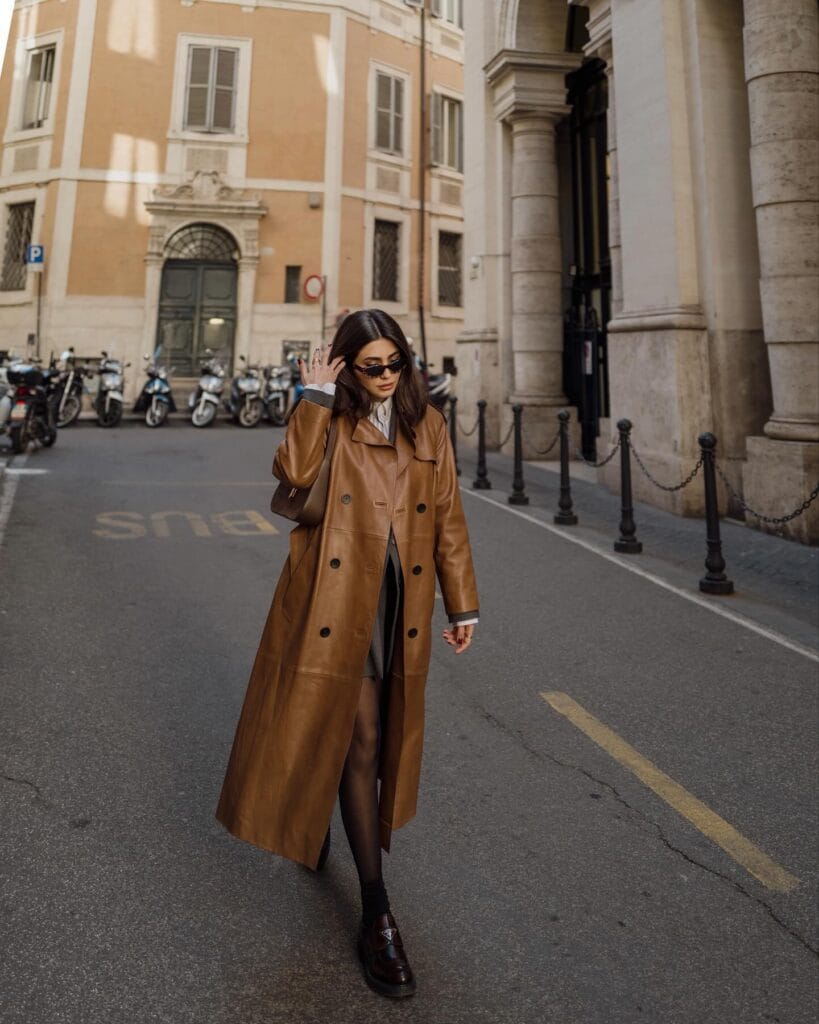 Woman wearing a long tan leather trench coat, Floor-Sweeping Coat trend, city street winter style.
