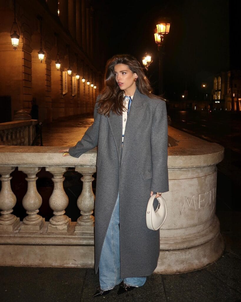 Model in oversized gray coat and jeans at night, Floor-Sweeping Coat winter trend, city lights background.

