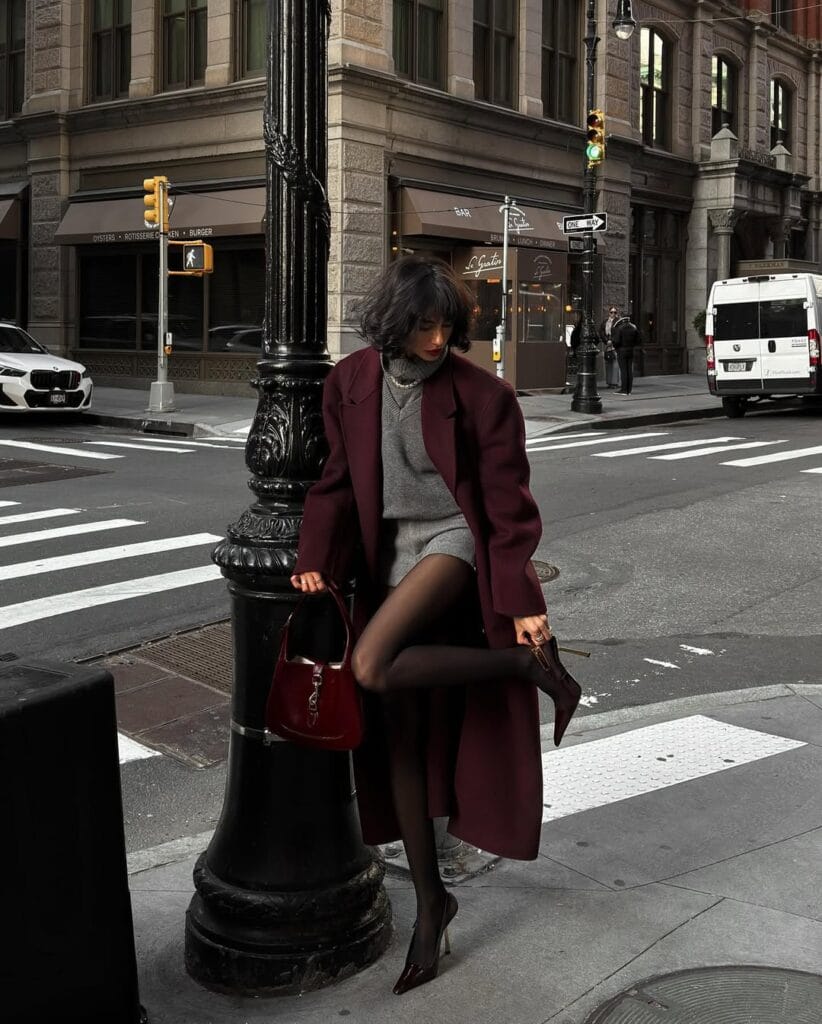 Long burgundy coat over gray layers, Floor-Sweeping Coat trend, winter street style with heels and tights.
