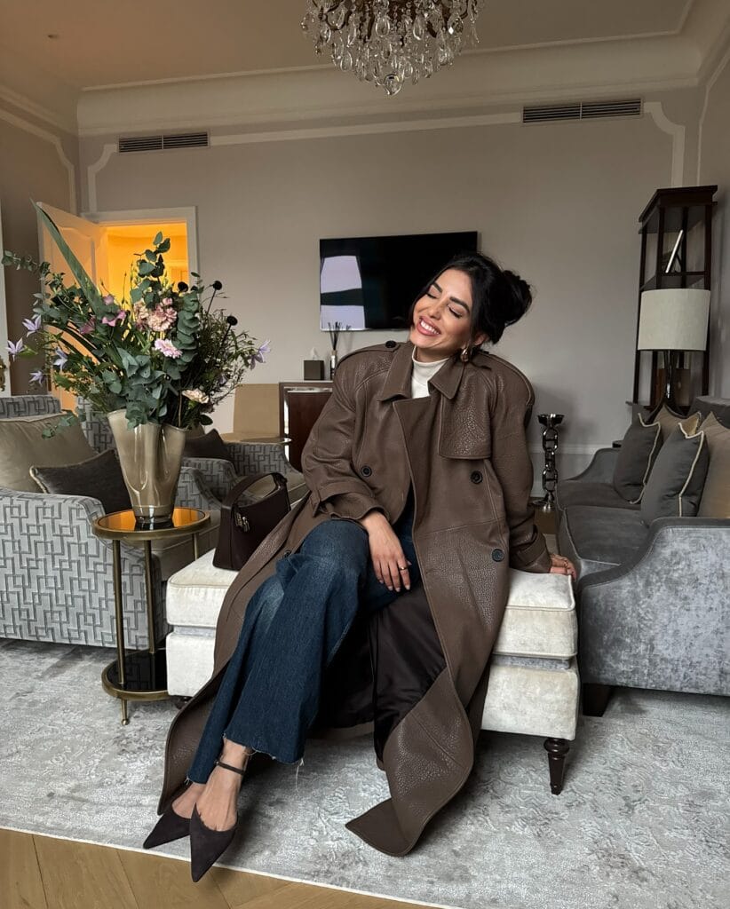 woman in brown trench coat, wide-leg cropped jeans, heels, sitting in elegant living room
