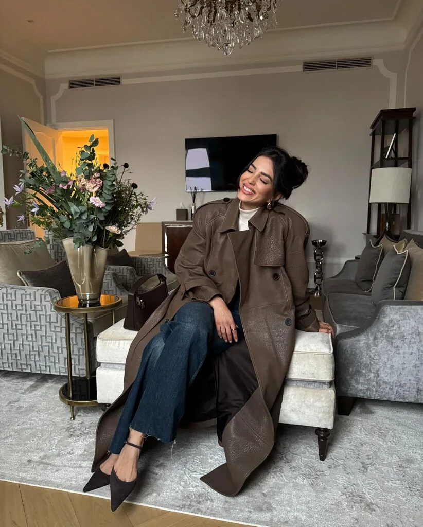 woman in brown trench coat, wide-leg cropped jeans, heels, sitting in elegant living room