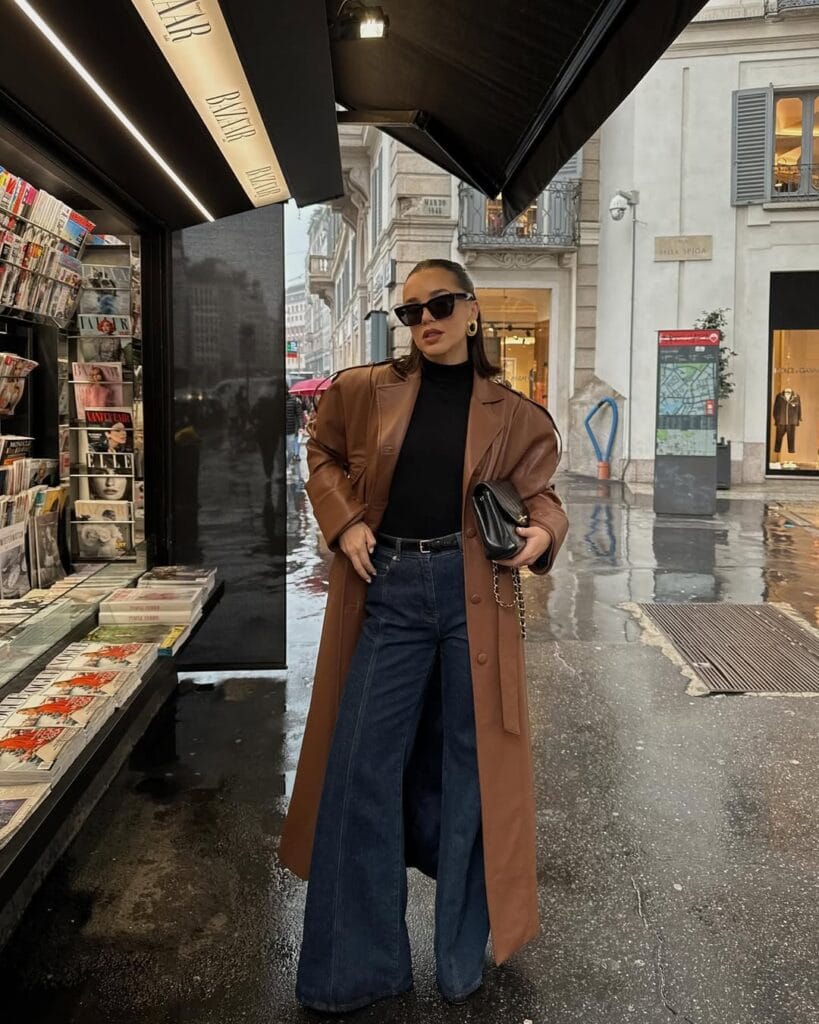 woman in chestnut leather coat, black turtleneck, wide-leg dark jeans, black clutch, rainy street look
