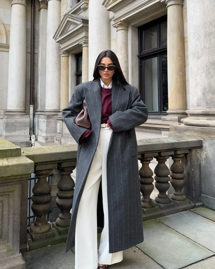winter work outfit with charcoal pinstripe long coat, burgundy sweater over white shirt, white trousers, sunglasses, clutch
