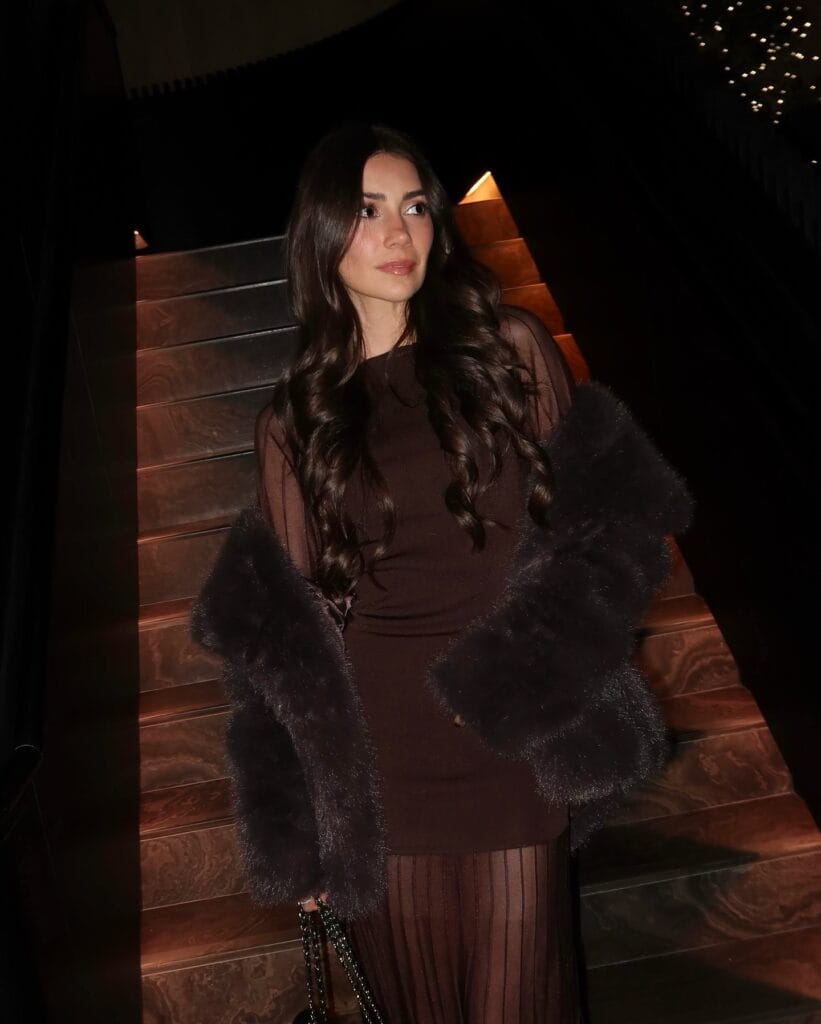 Woman in sheer brown dress with fur stole on dim staircase, elegant holiday outfit.
