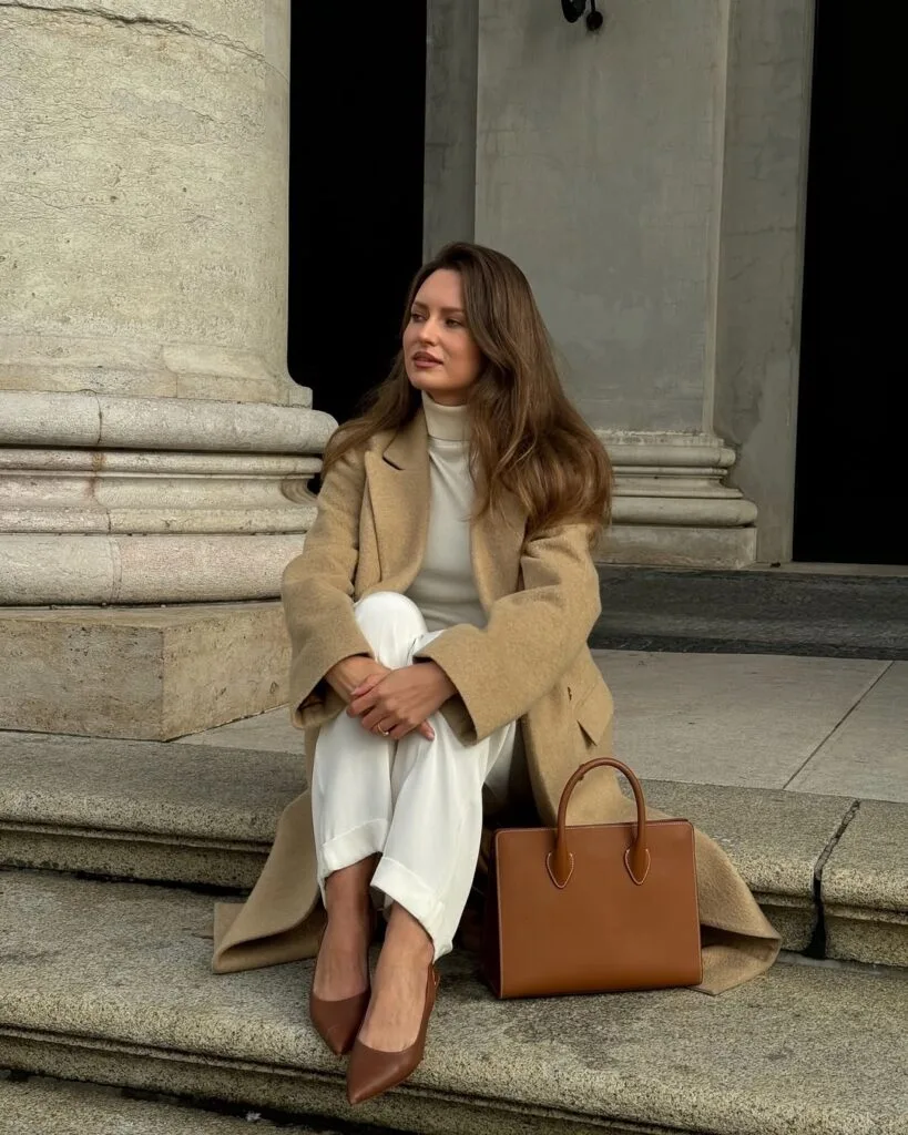 woman wearing camel coat, white turtleneck, white trousers, brown pumps, brown handbag, minimalist winter outfit