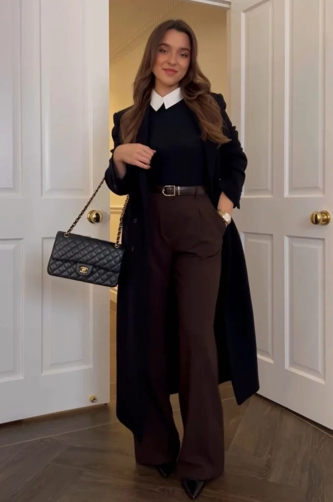 Woman in dark navy long coat over black sweater with white collar, brown trousers, and pointed-toe heels.
