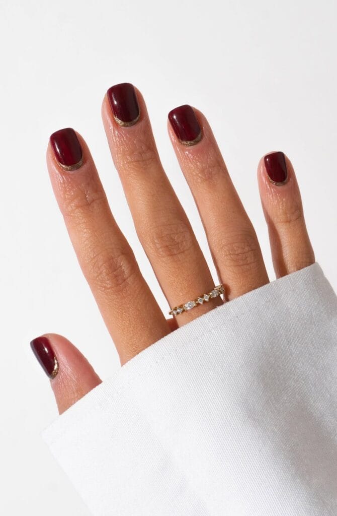 short winter nails in burgundy with thin gold glitter reverse French at cuticle, square shape, silver ring