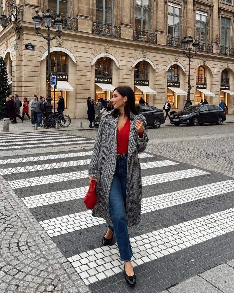 woman wearing herringbone coat, red knit sweater, blue jeans, black ballet flats, red bag, chic winter outfit
