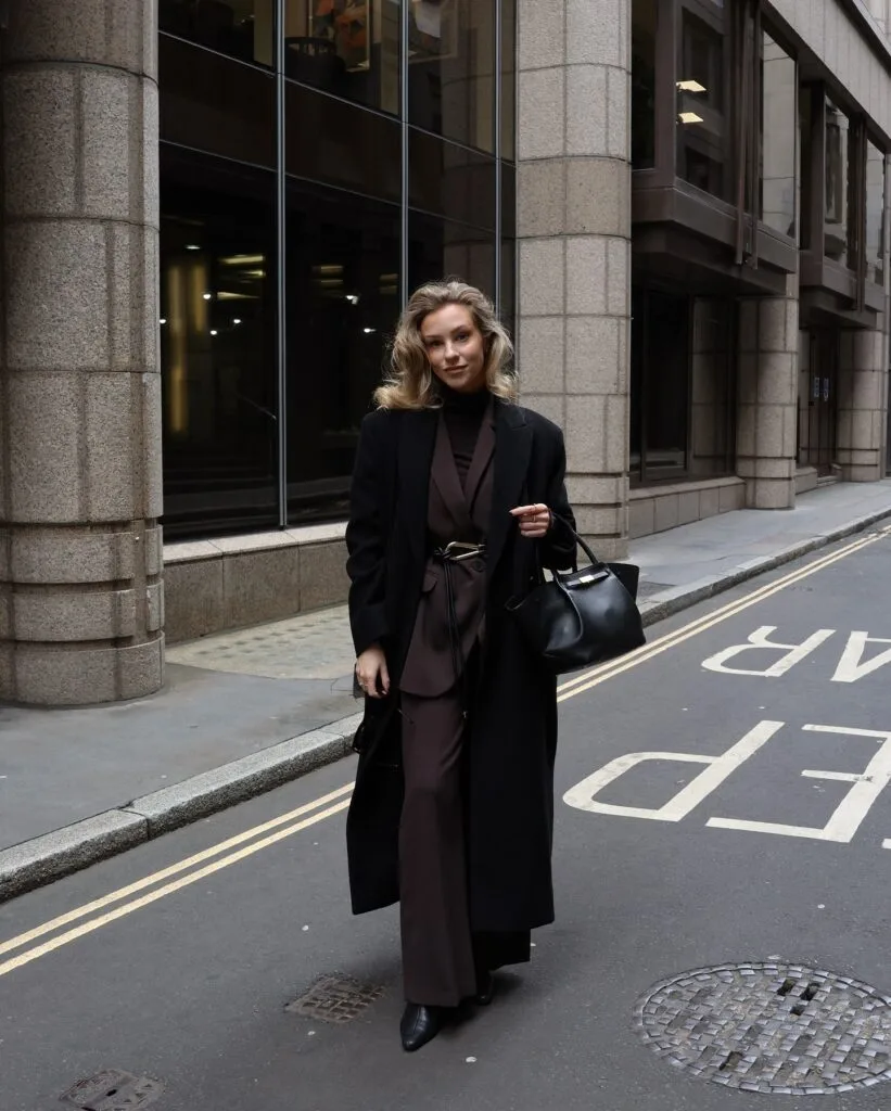 Woman in black coat over brown suit and black turtleneck, carrying a black handbag on a city street.
