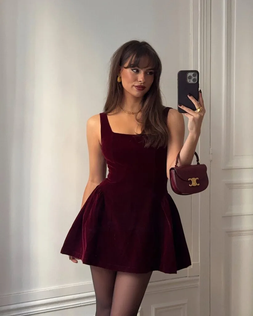 Woman in burgundy velvet mini dress with gold earrings and small burgundy bag, New Year’s Eve outfit.

