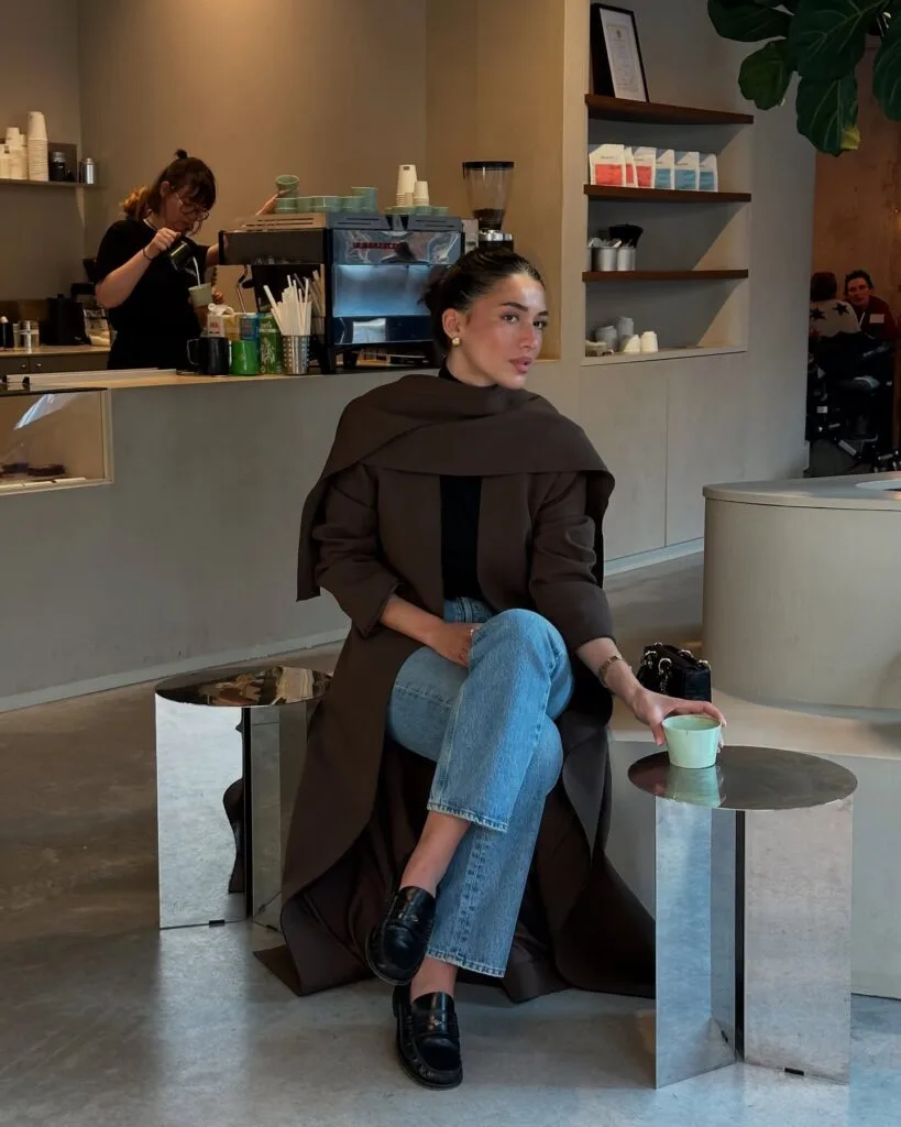 woman in brown wool coat, jeans, black loafers, and scarf, sitting outside at a café, winter style