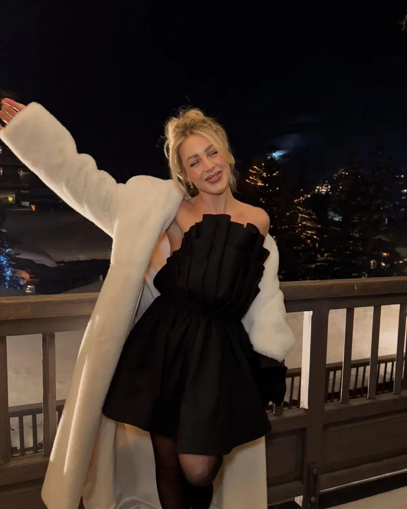 Woman in black sculpted mini dress and white faux fur coat outdoors at night, New Year’s Eve outfit.
