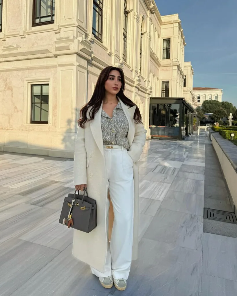 Cream coat with patterned blouse and white trousers, styled with sneakers and a structured gray bag for winter work.
